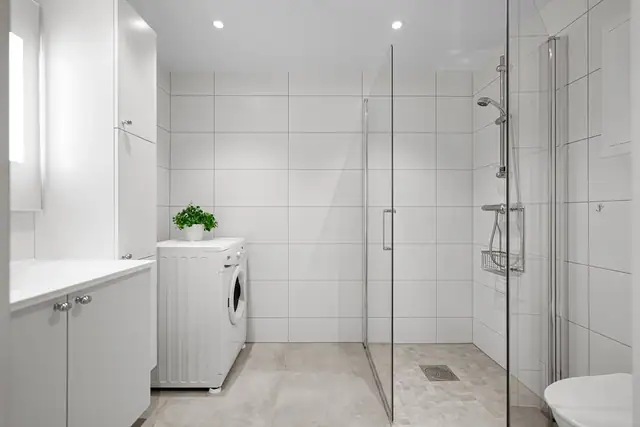 Flott, lyst og flislagt bad med glass dusjvegger, vegghengt toalett og opplegg for vaskemaskin.